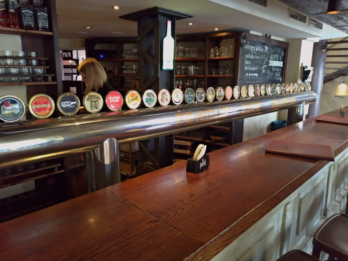 shutterstock_2608422763 1 | TVGURU.cz Row of beer tap handles along a wooden bar in a cozy pub interior with glassware on shelves behind it.