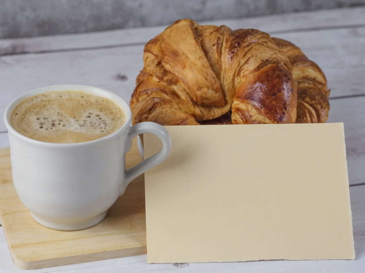 Na stole leží hrnek s kávou, kartička a croissant.