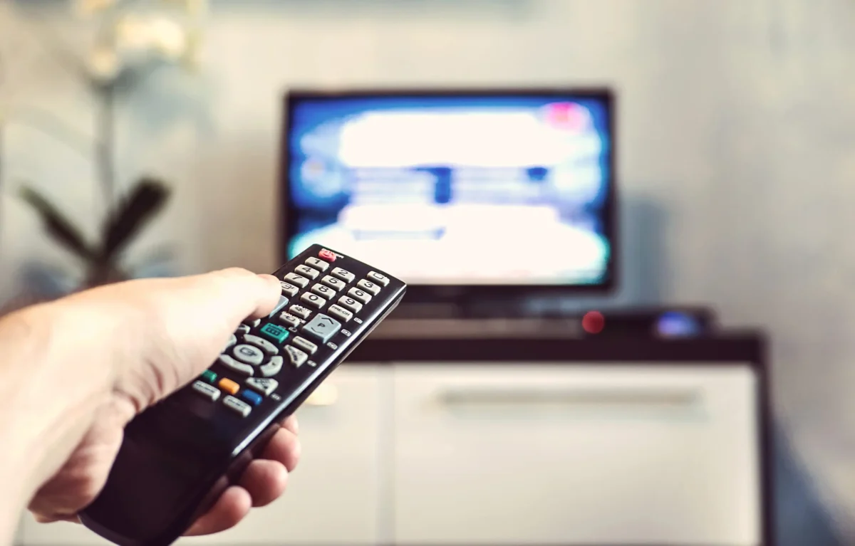 shutterstock_639613450 1 | TVGURU.cz Close-up of a hand holding a TV remote aimed at a blurred television screen in the background.
