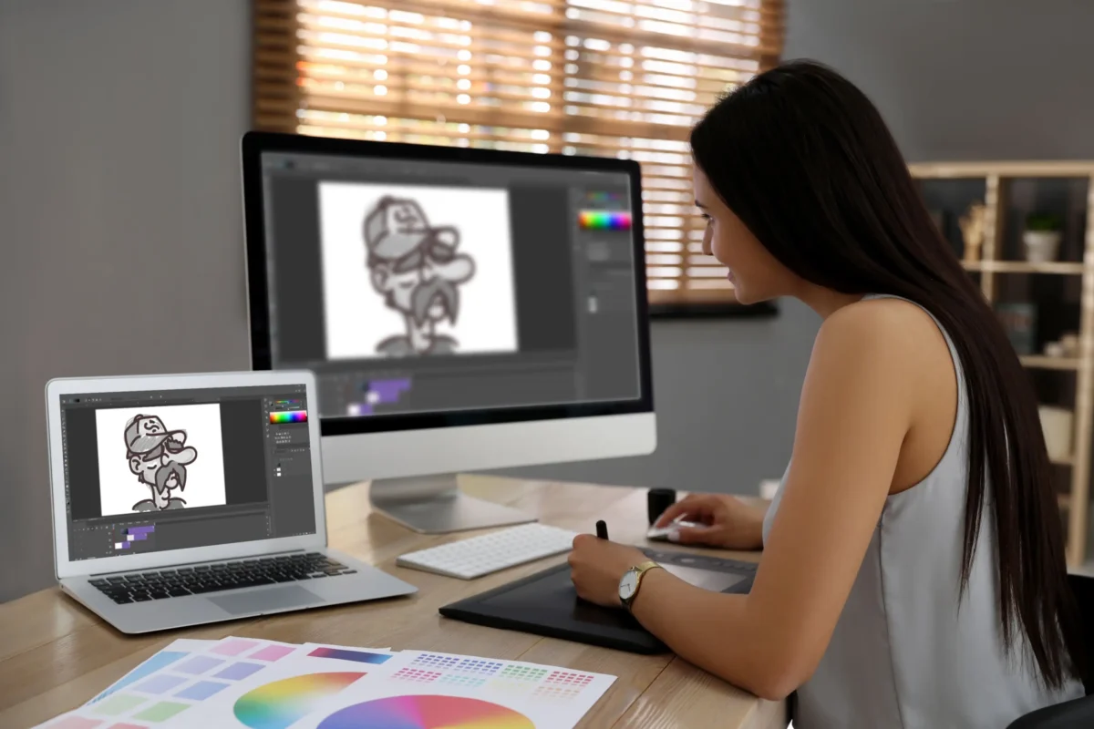 Designer at a desk drawing a cartoon on a desktop monitor, with a graphics tablet and color swatches nearby.