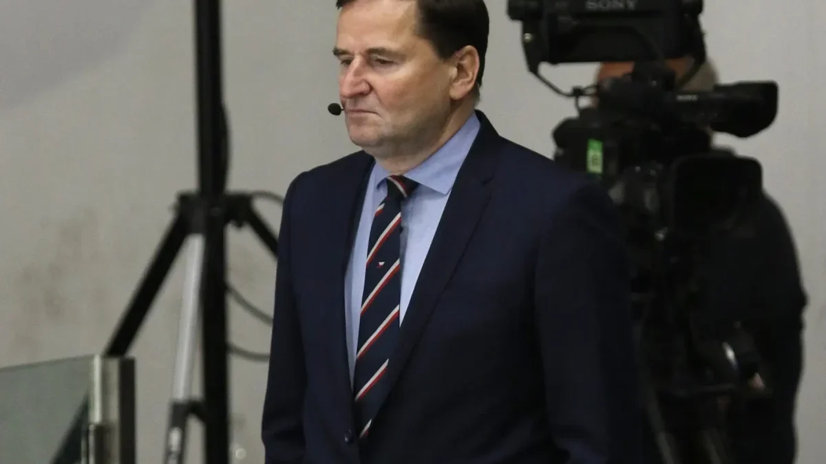 Man in a dark suit and striped tie with a headset mic, standing in front of TV cameras in a studio or newsroom setting.