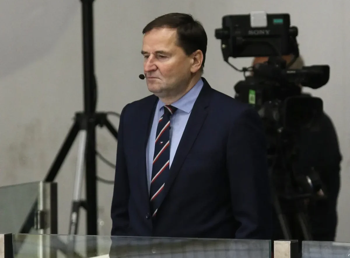 Man in a dark suit and striped tie with a headset mic, standing in front of TV cameras in a studio or newsroom setting.