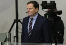 Man in a dark suit and striped tie with a headset mic, standing in front of TV cameras in a studio or newsroom setting.