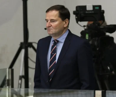 Man in a dark suit and striped tie with a headset mic, standing in front of TV cameras in a studio or newsroom setting.