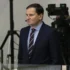 Man in a dark suit and striped tie with a headset mic, standing in front of TV cameras in a studio or newsroom setting.
