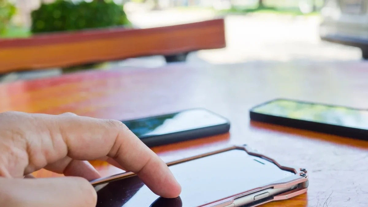 Close-up of a hand tapping a touchscreen smartphone on a wooden table, with other devices blurred in the background.
