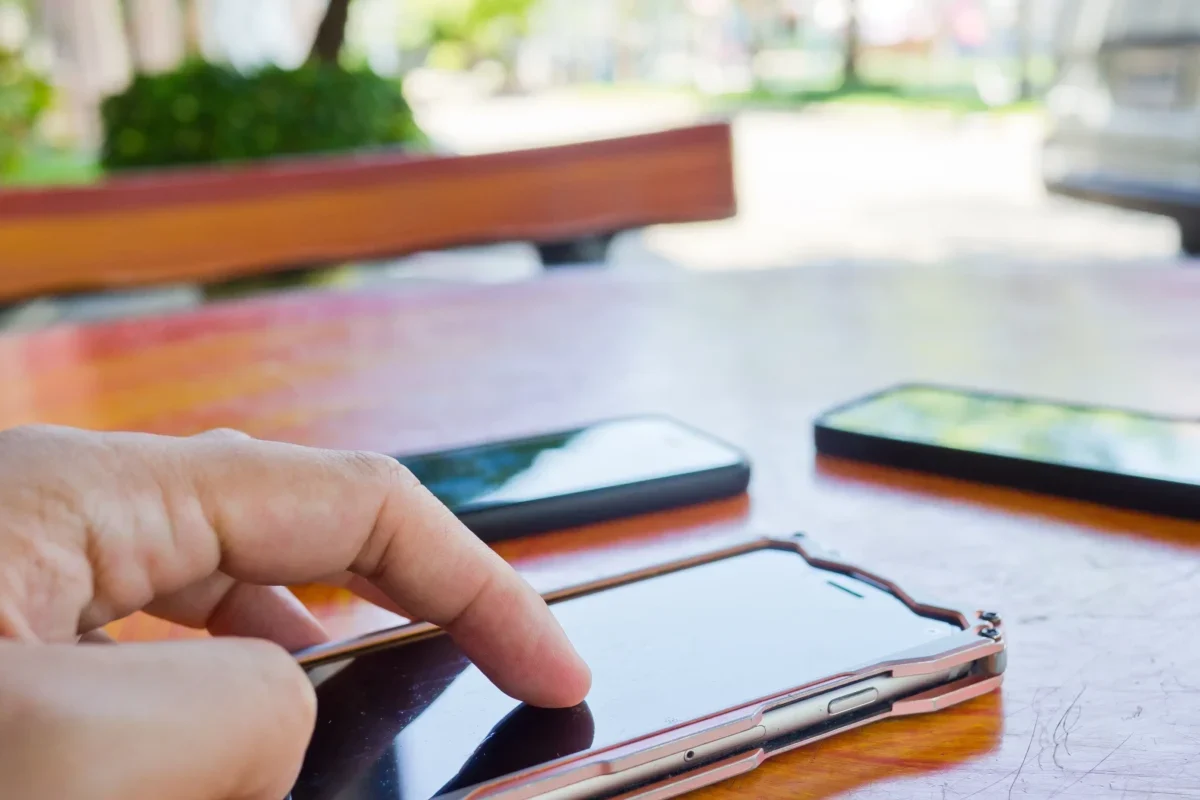 Close-up of a hand tapping a touchscreen smartphone on a wooden table, with other devices blurred in the background.