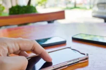 Close-up of a hand tapping a touchscreen smartphone on a wooden table, with other devices blurred in the background.
