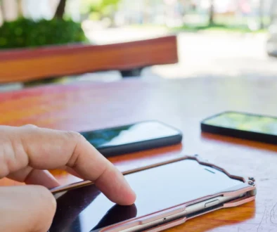 Close-up of a hand tapping a touchscreen smartphone on a wooden table, with other devices blurred in the background.