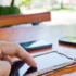 Close-up of a hand tapping a touchscreen smartphone on a wooden table, with other devices blurred in the background.