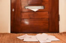 Wooden front door with a metal mail slot; a white envelope pokes out, papers scattered on the carpet in front of it.