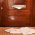 Wooden front door with a metal mail slot; a white envelope pokes out, papers scattered on the carpet in front of it.