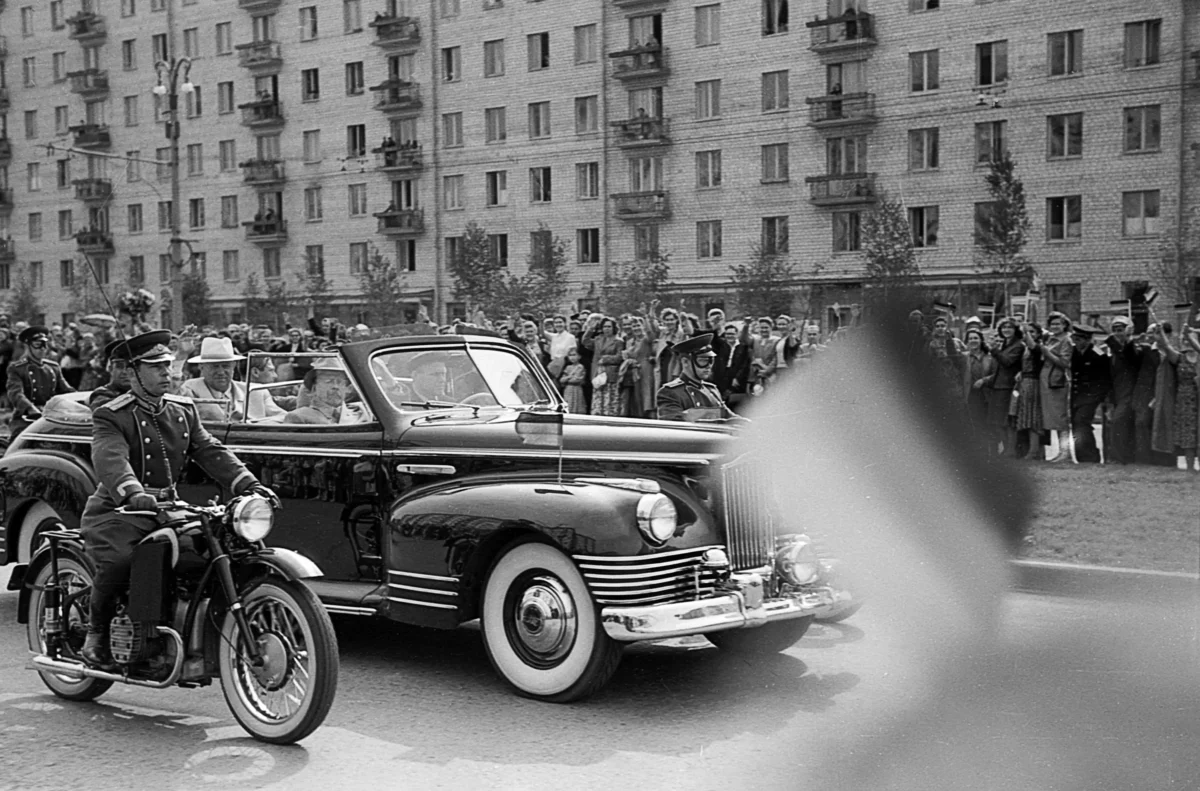 shutterstock_2672775063 | TVGURU.cz Na černobílé fotografii je historické auto a lidé.