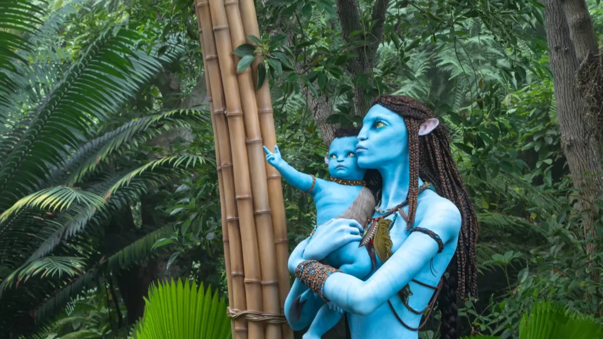 Life-sized blue Na’vi figures, a mother and child, touching a bamboo post in a dense jungle setting.