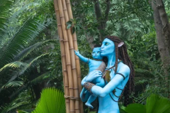Life-sized blue Na’vi figures, a mother and child, touching a bamboo post in a dense jungle setting.