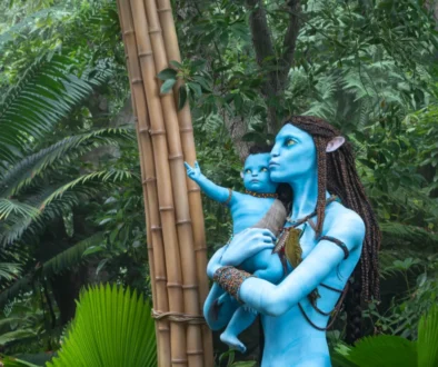 Life-sized blue Na’vi figures, a mother and child, touching a bamboo post in a dense jungle setting.