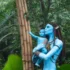 Life-sized blue Na’vi figures, a mother and child, touching a bamboo post in a dense jungle setting.