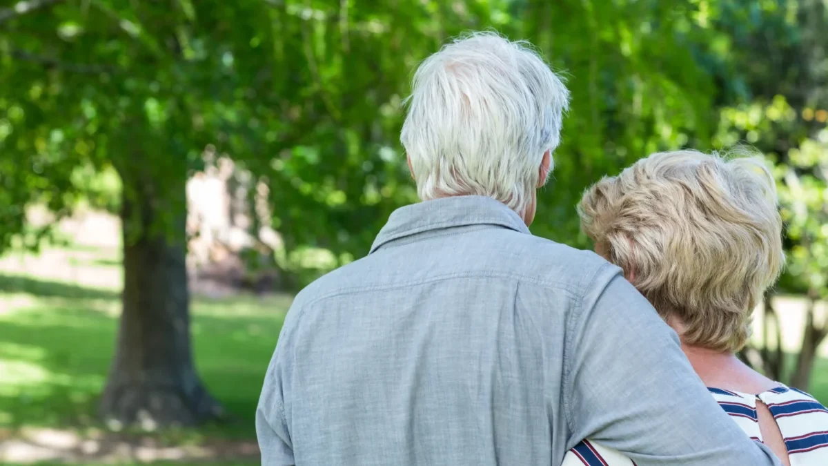 Two older adults seen from behind, embracing in a sunny park with green trees in the background