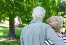 Two older adults seen from behind, embracing in a sunny park with green trees in the background