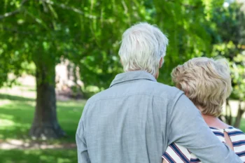 Two older adults seen from behind, embracing in a sunny park with green trees in the background