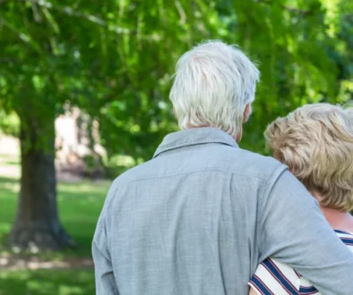 Two older adults seen from behind, embracing in a sunny park with green trees in the background