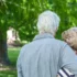 Two older adults seen from behind, embracing in a sunny park with green trees in the background