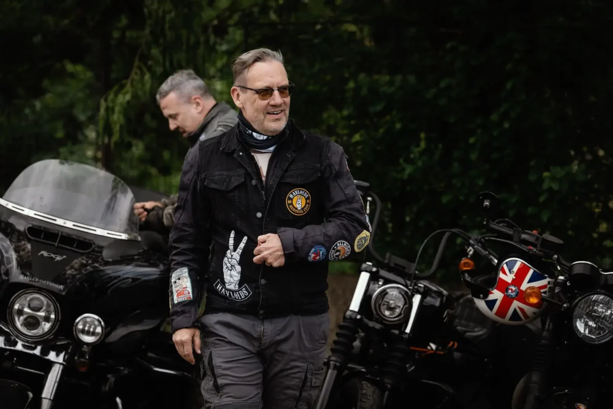 profimedia-1098103836 1 | TVGURU.cz Man with sunglasses and a patch-covered black jacket stands beside parked motorcycles in a green outdoor area, smiling slightly.