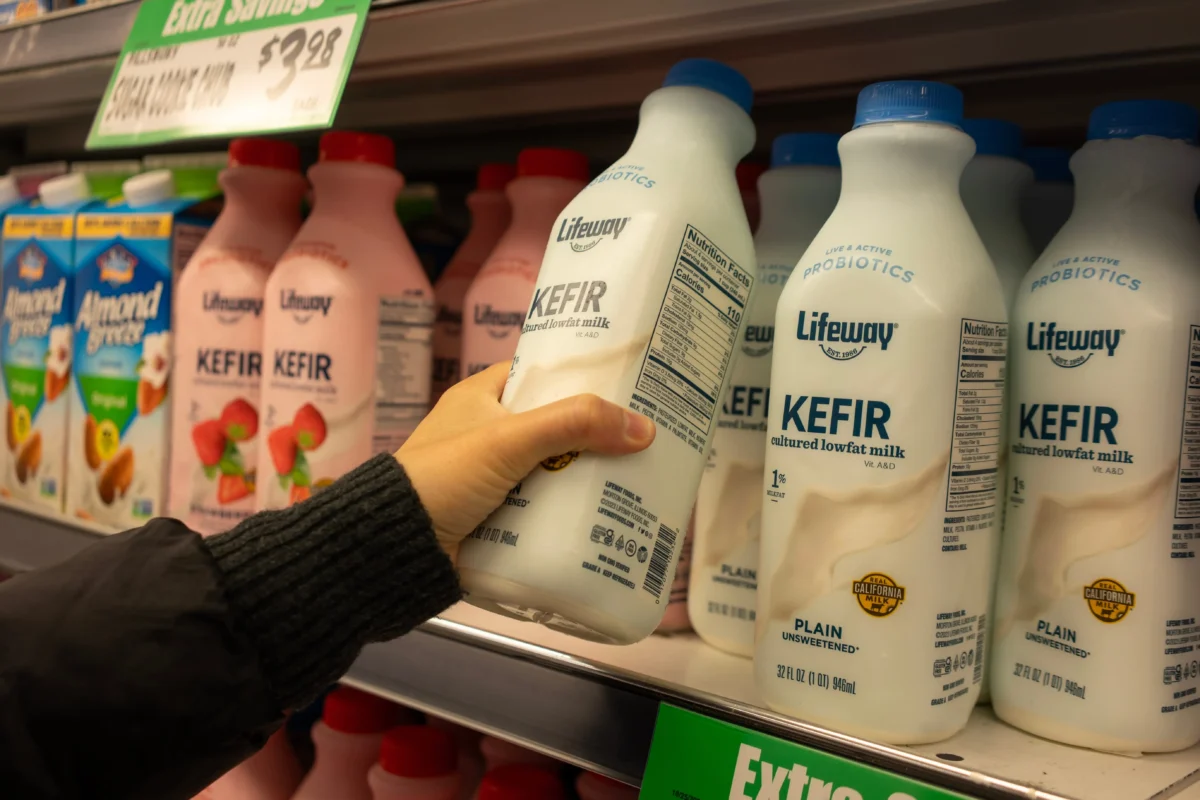 shutterstock_2517796681 1 | TVGURU.cz Person reaching to grab Lifeway kefir bottles from a dairy shelf in a grocery store.