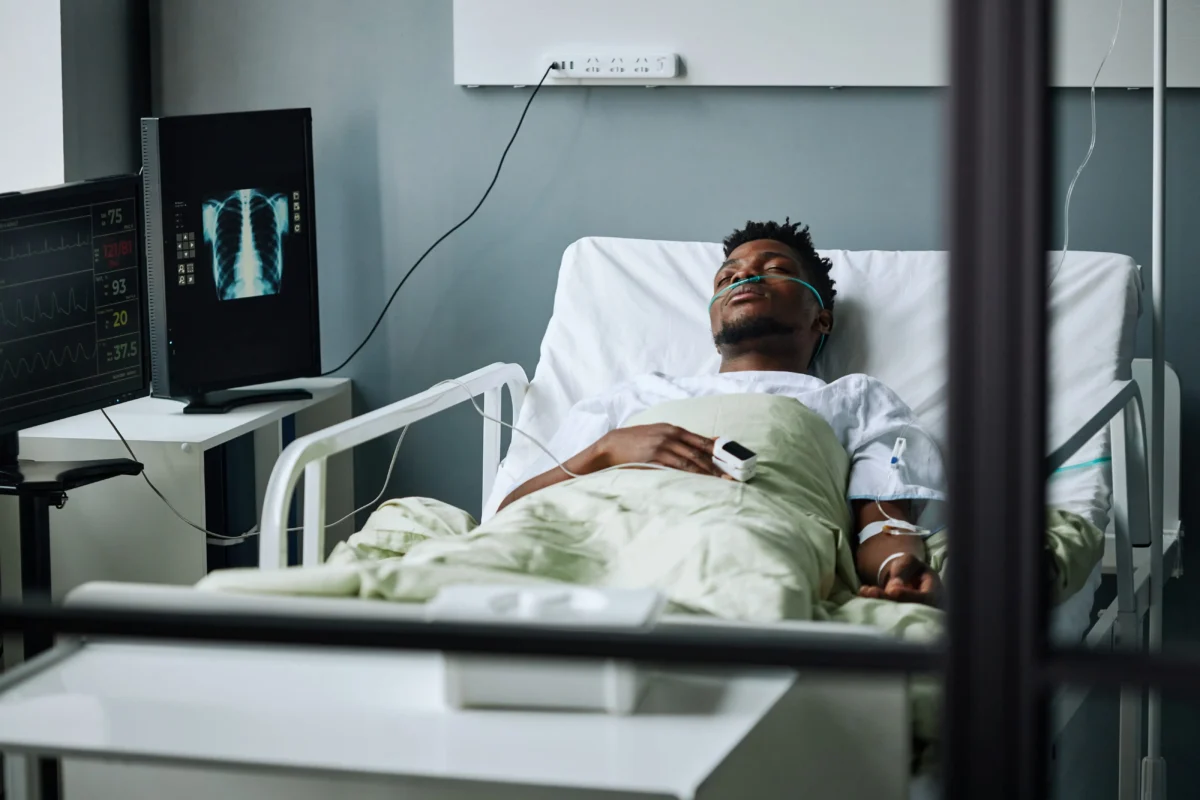 shutterstock_2708694699 1 | TVGURU.cz Patient resting in a hospital bed with oxygen tubing, surrounded by monitoring screens and a medical X-ray image on a nearby monitor.