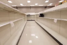 Empty supermarket aisle with bare shelves on both sides and bright ceiling lights.