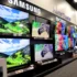 Showroom wall with large flat-screen TVs displaying colorful landscapes and product brands like Samsung, Sony, and LG