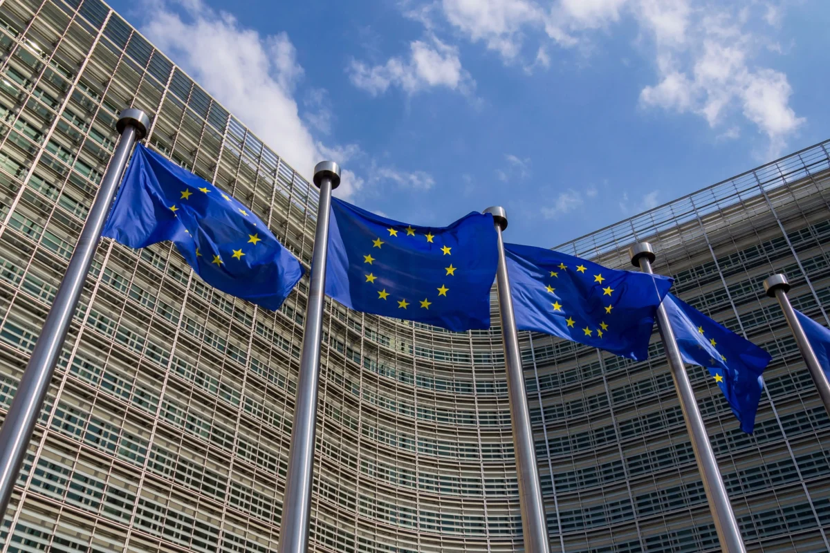 EU flags fluttering in front of a modern glass office building against a blue sky.