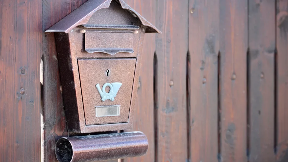 Rusty brown wall-mounted mailbox with a small roof, affixed to a weathered wooden fence.