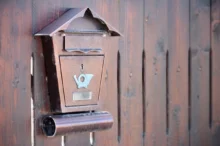 Rusty brown wall-mounted mailbox with a small roof, affixed to a weathered wooden fence.