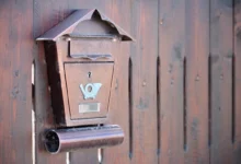 Rusty brown wall-mounted mailbox with a small roof, affixed to a weathered wooden fence.