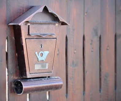Rusty brown wall-mounted mailbox with a small roof, affixed to a weathered wooden fence.
