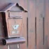 Rusty brown wall-mounted mailbox with a small roof, affixed to a weathered wooden fence.