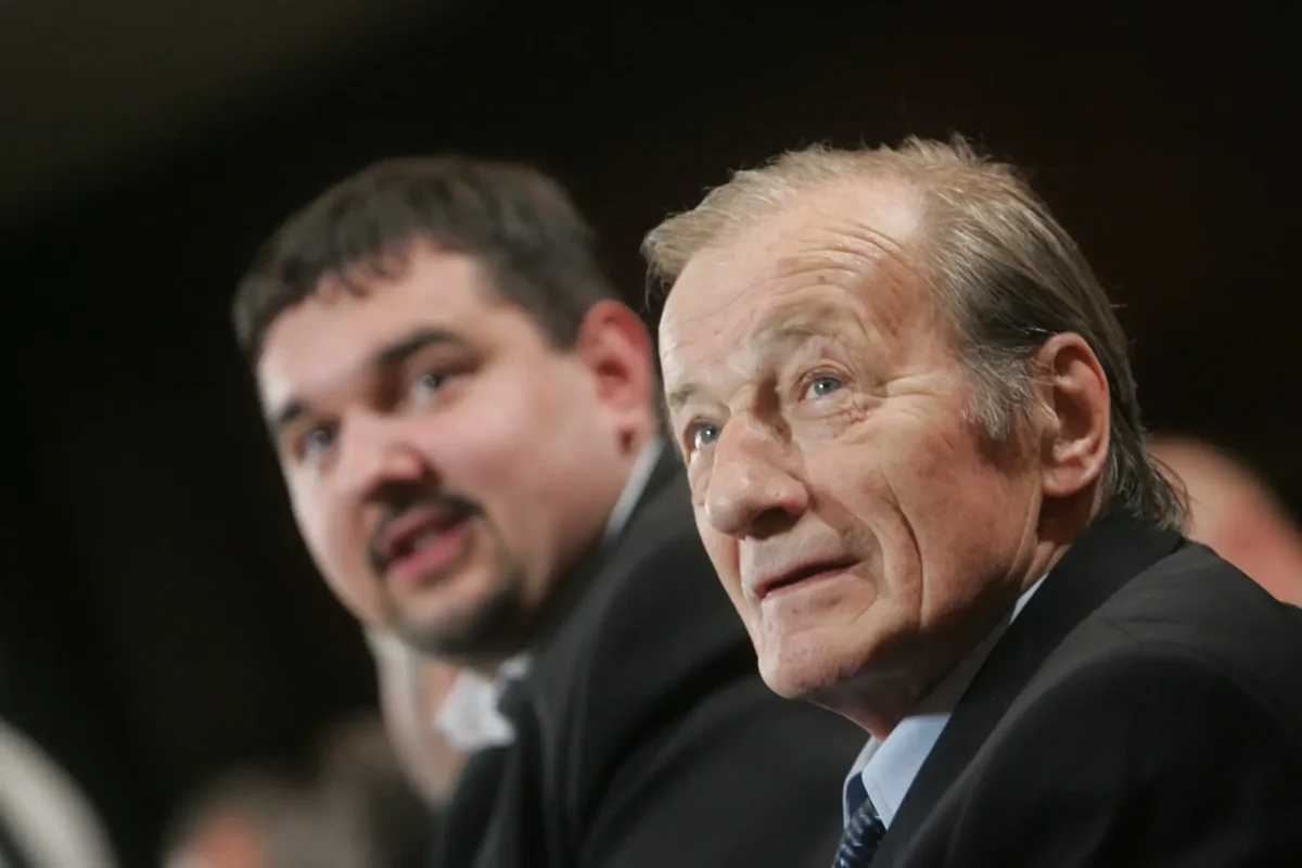 Close-up of an older man in a suit looking upward, with a younger man blurred in the background at a formal event.