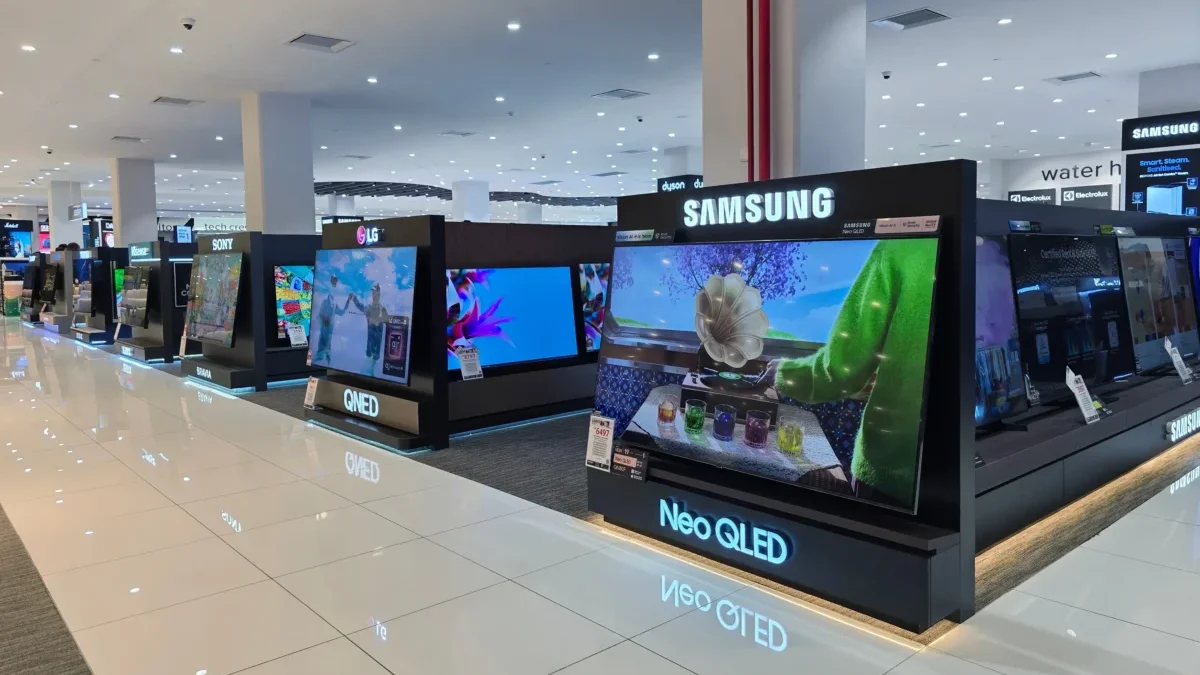 Bright electronics store with rows of TV displays on black platforms, Samsung signage overhead, polished white tiled floor and overhead lights reflecting on the floor.