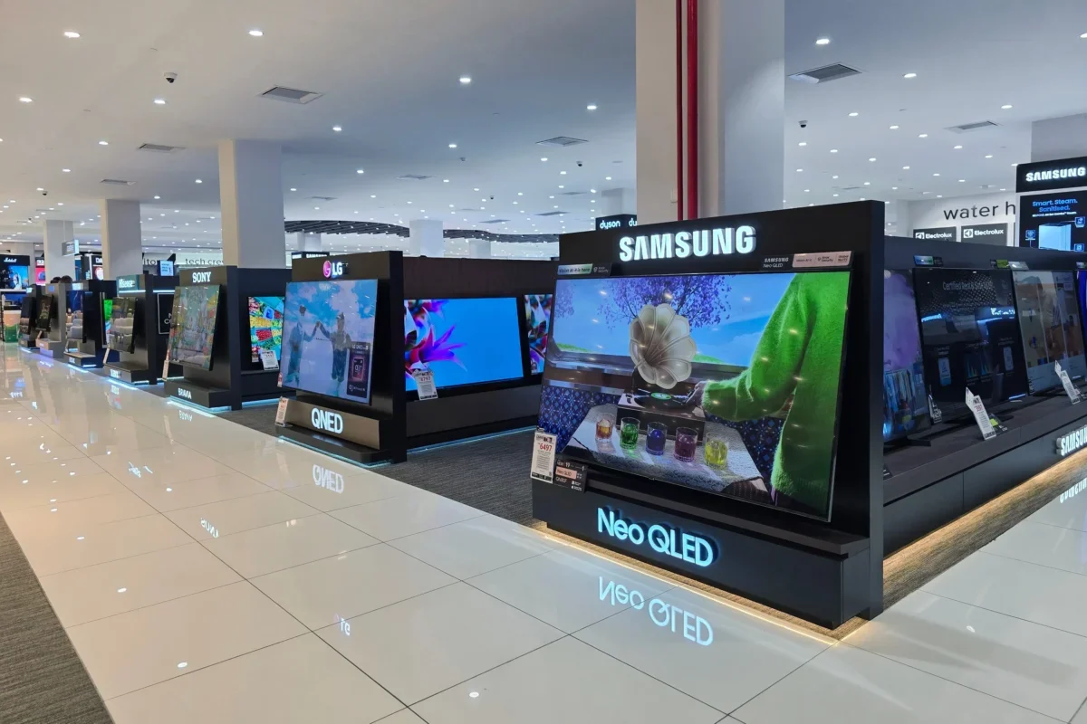Bright electronics store with rows of TV displays on black platforms, Samsung signage overhead, polished white tiled floor and overhead lights reflecting on the floor.