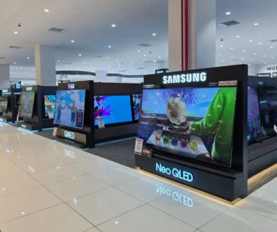 Bright electronics store with rows of TV displays on black platforms, Samsung signage overhead, polished white tiled floor and overhead lights reflecting on the floor.