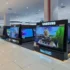 Bright electronics store with rows of TV displays on black platforms, Samsung signage overhead, polished white tiled floor and overhead lights reflecting on the floor.