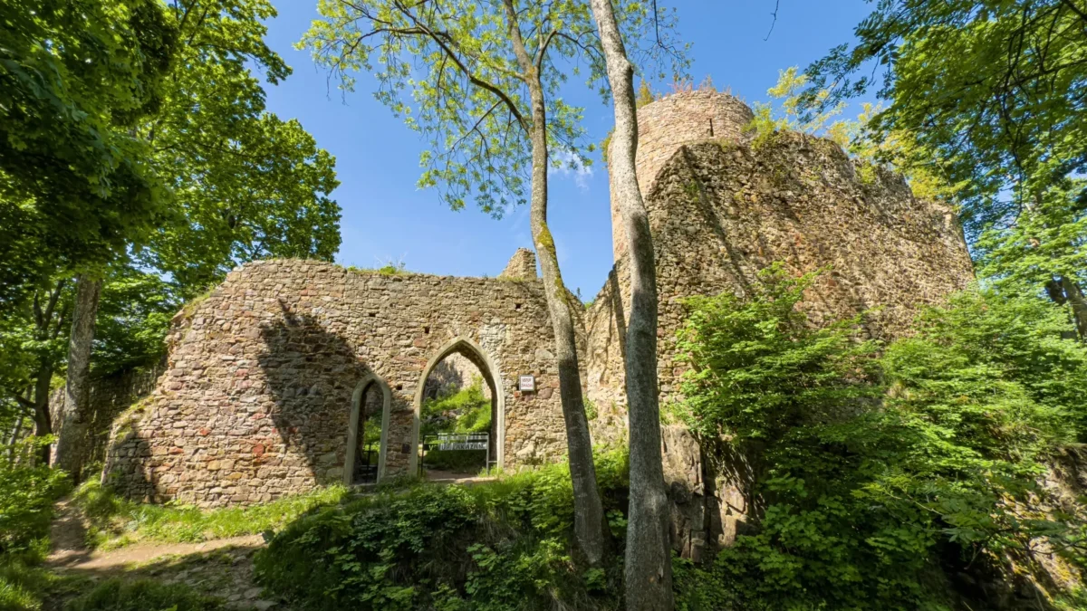 Old stone ruin with arched doorway, surrounded by dense greenery and trees under a bright blue sky