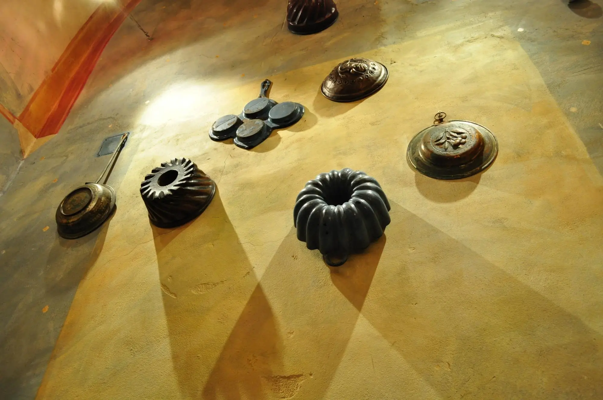 Domácí sbírka starožitného nádobí | TVGURU.cz Industrial metal objects arranged on a dusty floor, including circular vents, domed lids, and a ring-like sculpture, casting long shadows.