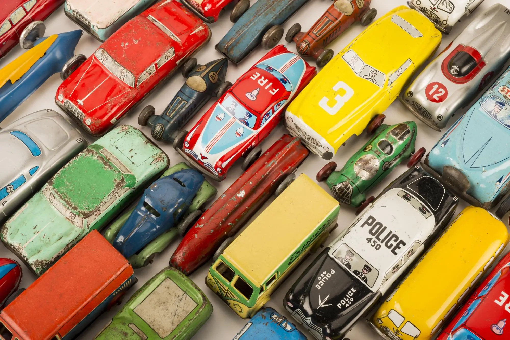 Sběratelé mají zájem o stará autíčka všeho druhu | TVGURU.cz Assorted vintage tin toy cars arranged diagonally, colorful and worn from play. 80s-era miniature vehicles in red, yellow, blue, and green.