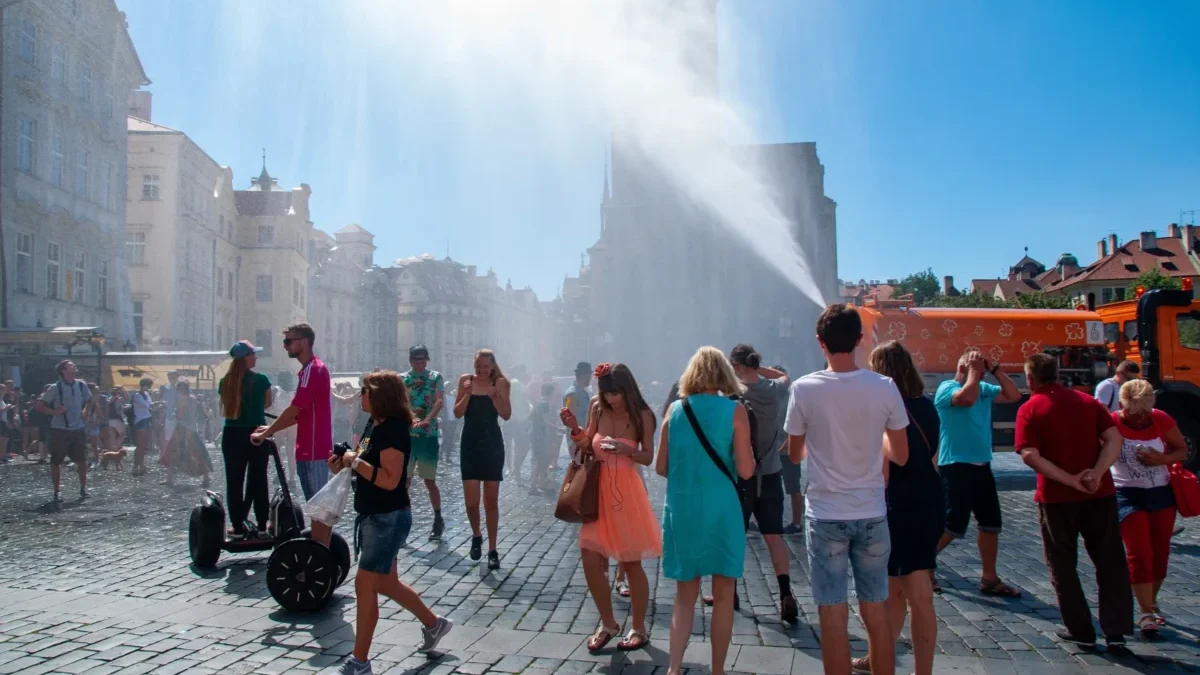 Crowd of people in a sunny cobblestone square as water jets spray across; historic buildings line the street behind them.