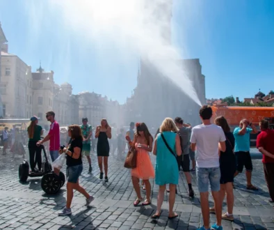 Crowd of people in a sunny cobblestone square as water jets spray across; historic buildings line the street behind them.