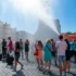 Crowd of people in a sunny cobblestone square as water jets spray across; historic buildings line the street behind them.