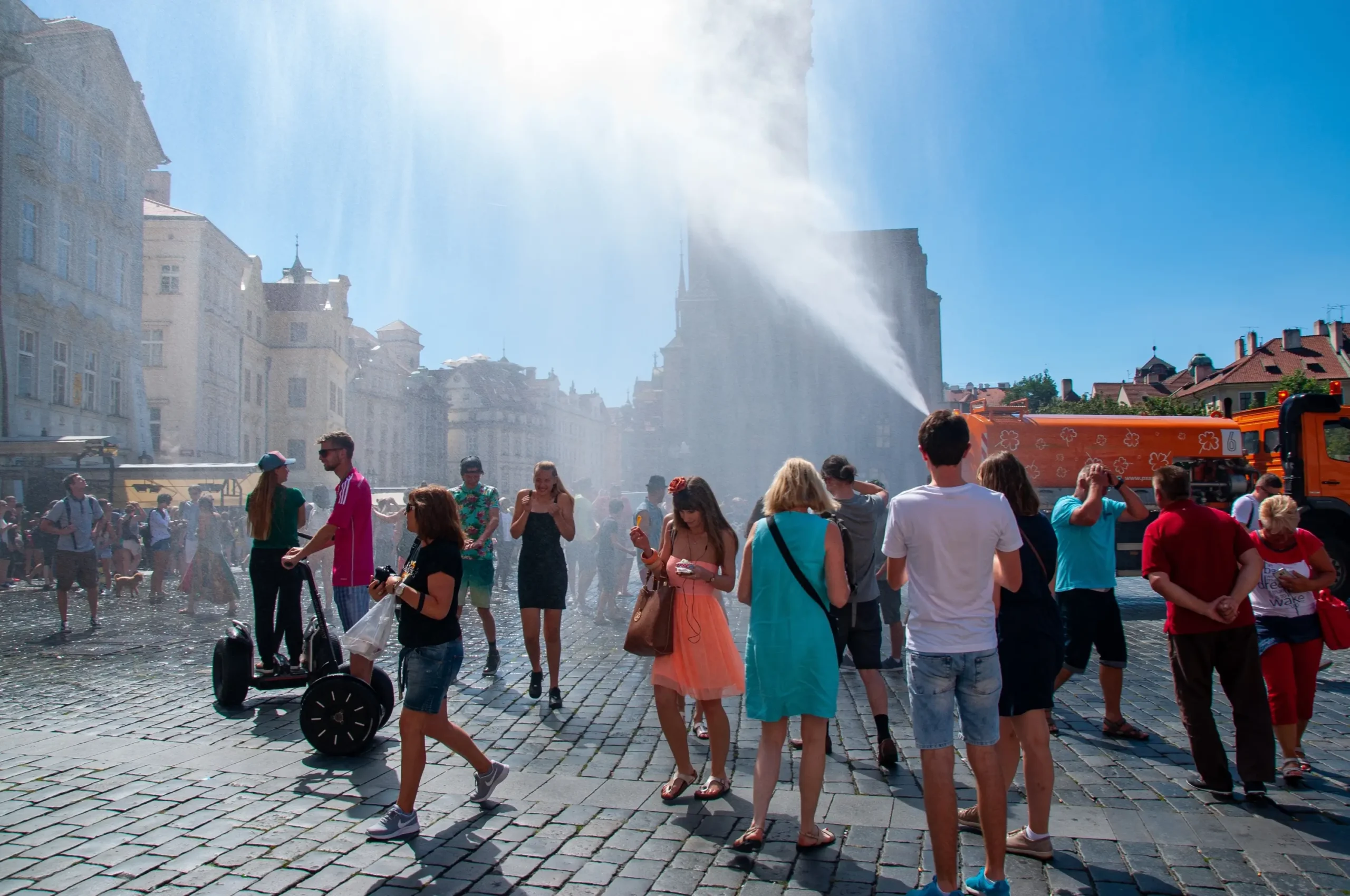 Crowd of people in a sunny cobblestone square as water jets spray across; historic buildings line the street behind them.