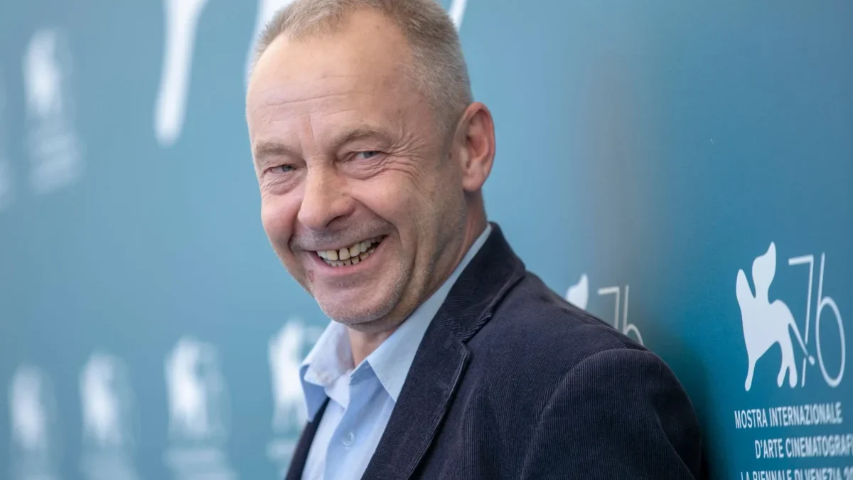 Smiling man in a suit posing against a blue wall with event signage in the background on the right side.